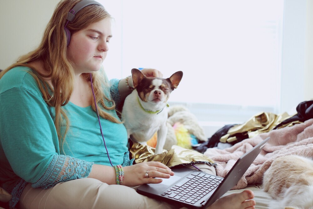 A girl on the bed with her dog, using a laptop with headphones