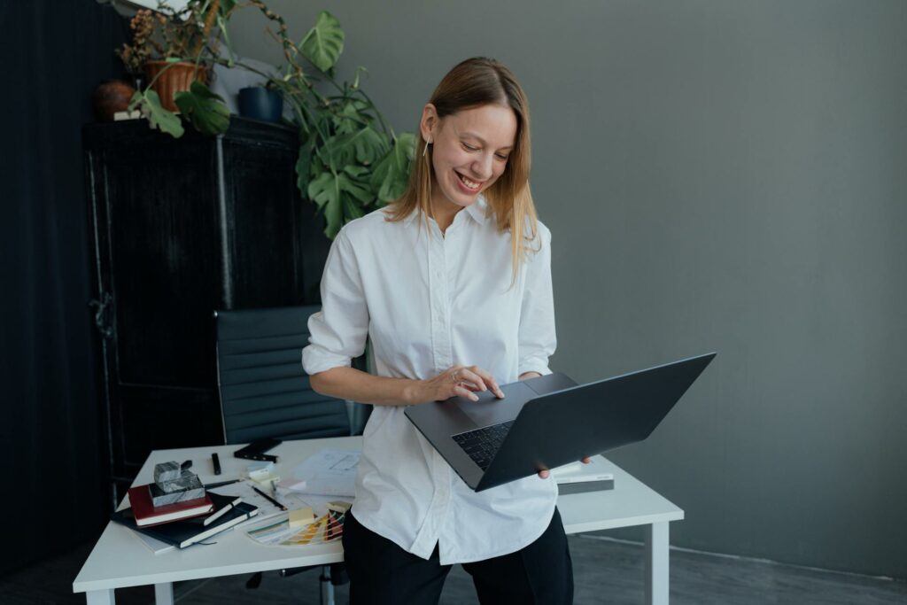 A woman leaning on a desk, looking at a laptop, with a pleased expression.
