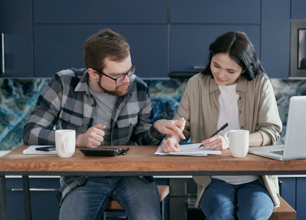 Two people at home, working through a problem with a calculator, a laptop, and notepad
